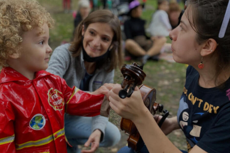 a young kid in a firefighter costume touching a violin that a performer is holding