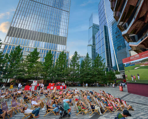 group of people sitting on the plaza watching golf on the Big Screen